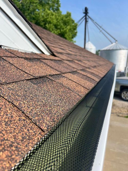 Close-up of brown shingle roof edge with black gutter guard beside silos and a driveway
