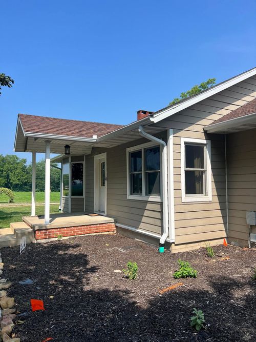 Small beige house exterior with white trim, porch columns, and a dark mulch garden under a blue sky