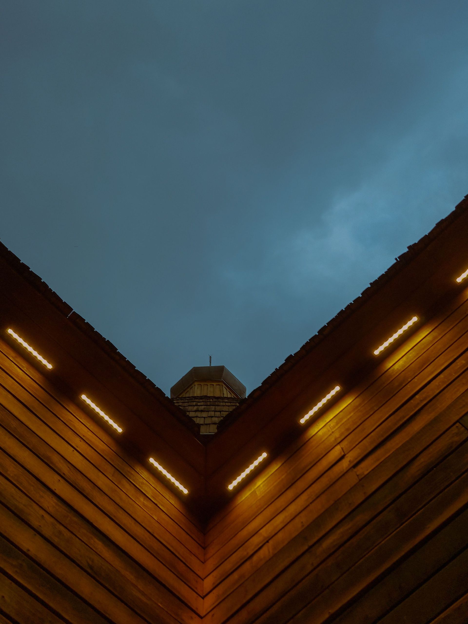 Symmetrical wooden V-shaped building corners with warm lights against a cloudy blue evening sky