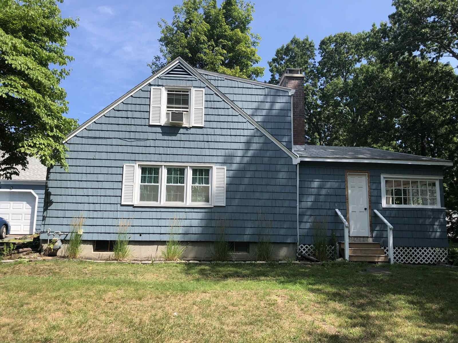 A blue, two-story house with white shutters, a brick chimney, and a small porch set against a backdrop of green trees.