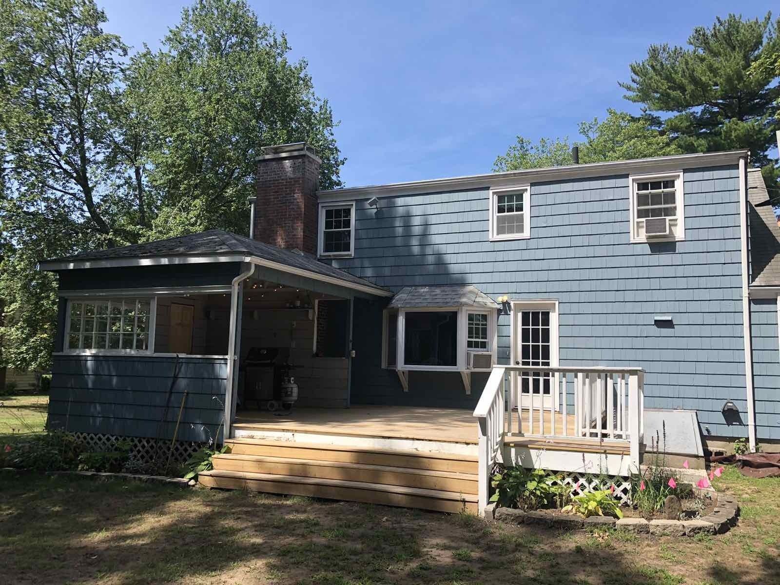 A two-story blue house with a wooden deck and a screened-in porch, set against a background of green trees and blue sky.