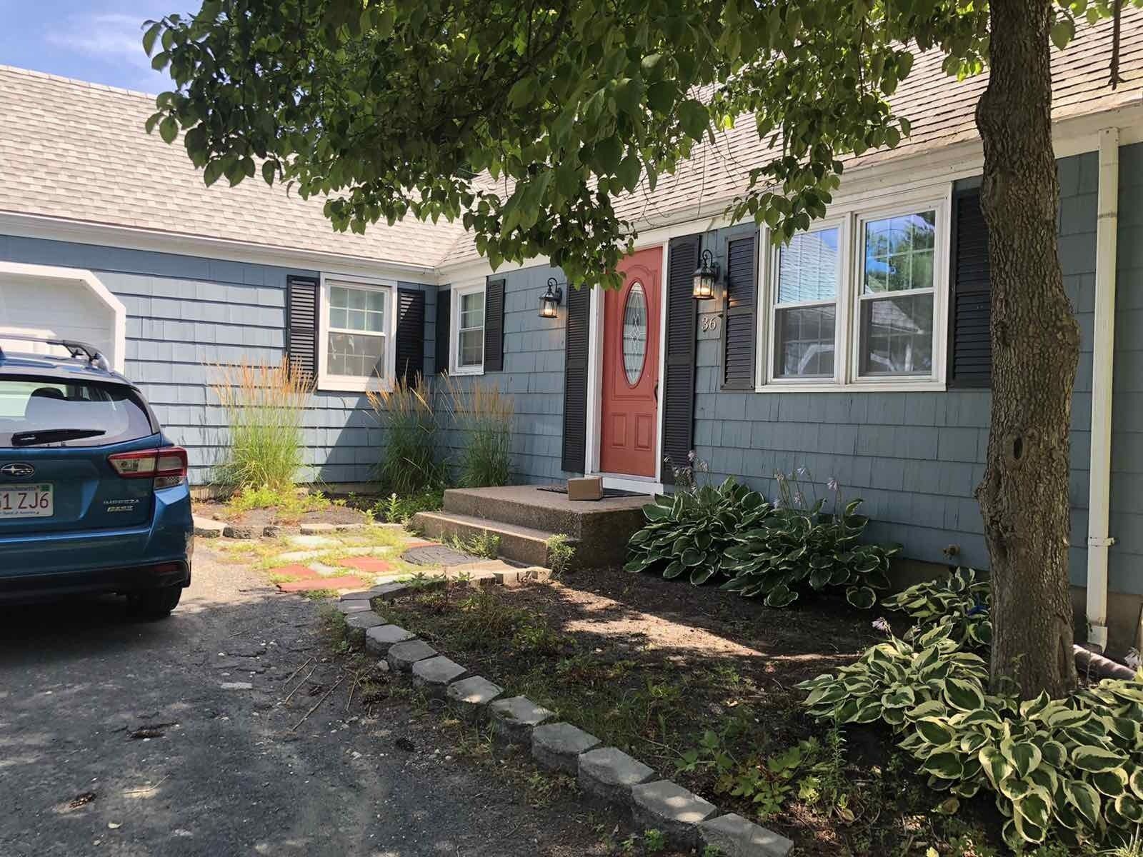 A blue house with a red front door, a driveway with a car parked on the left, and a landscaped garden in the front.