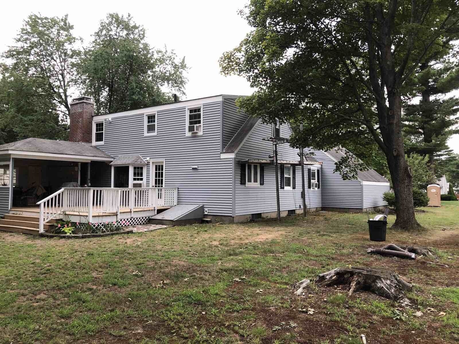 A grey-sided, two-story suburban house with a white deck, brick chimney, and a large tree in the front yard.