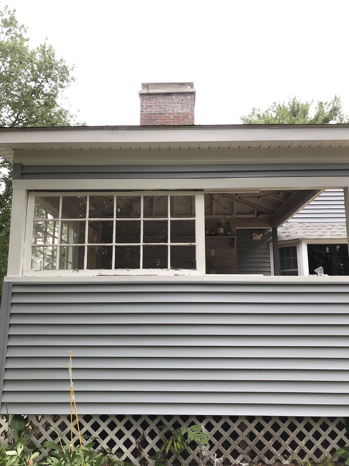 A gray-sided house exterior with a multi-pane window, white trim, a brick chimney, and lattice skirting below.