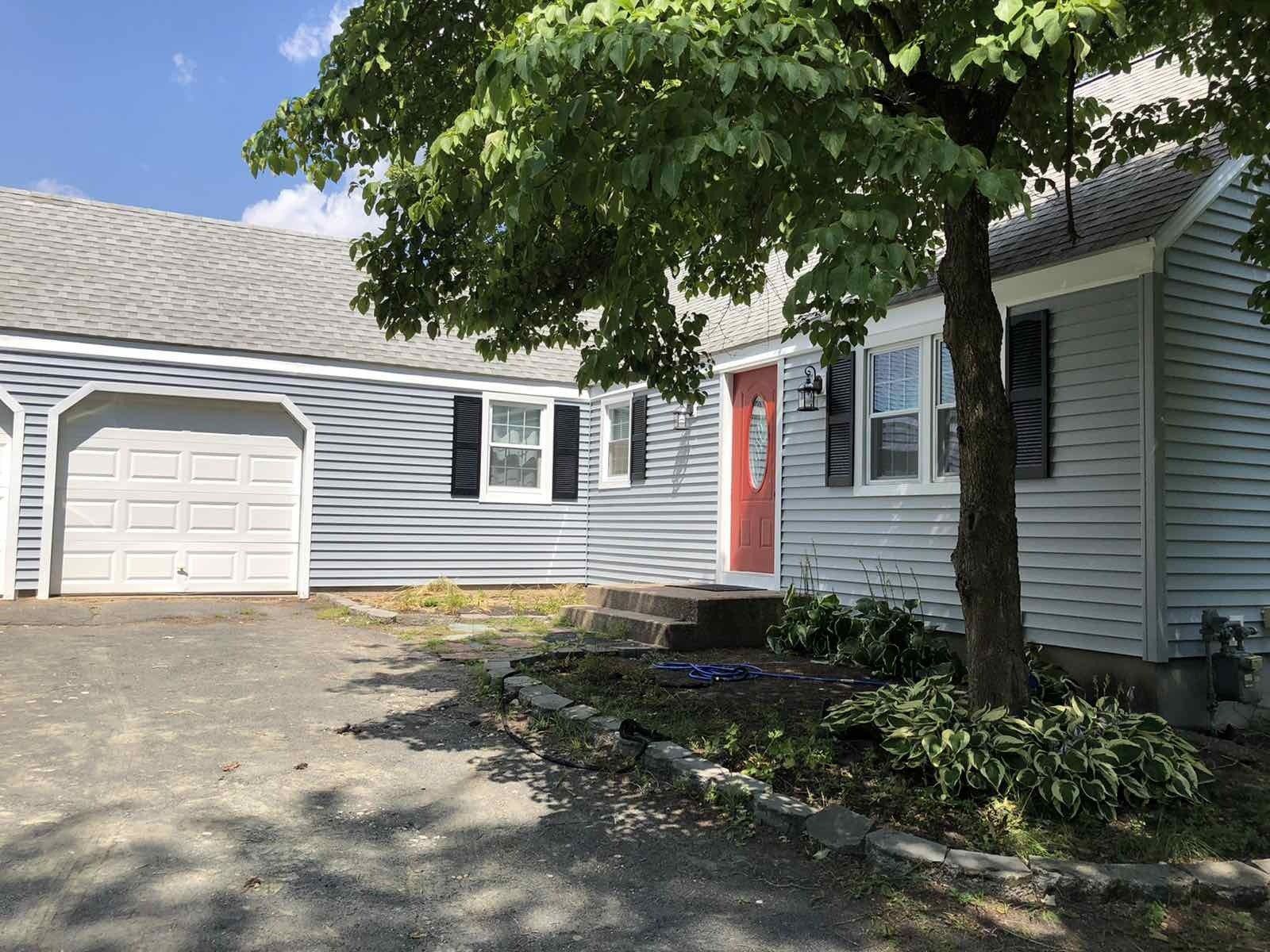 A gray, one-story house with a red front door, an attached garage, and a large tree in the front yard.