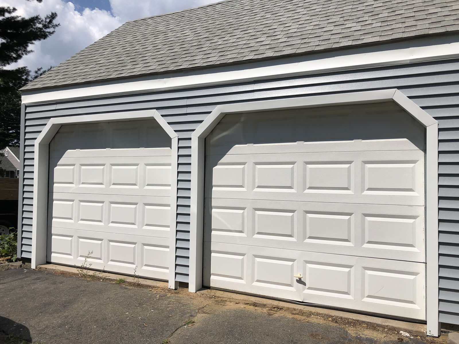 Two white, arched garage doors on a garage with grey horizontal siding under a shingled roof.