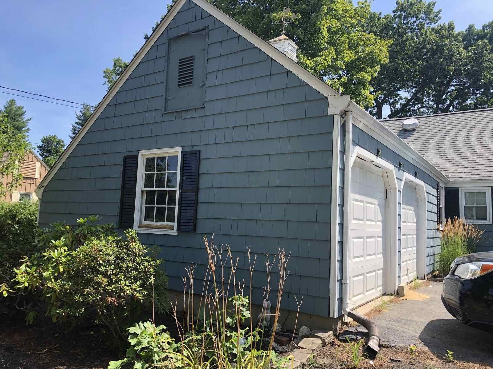 A blue garage with a white trim, a single window with black shutters, and a driveway with a car parked to the side.