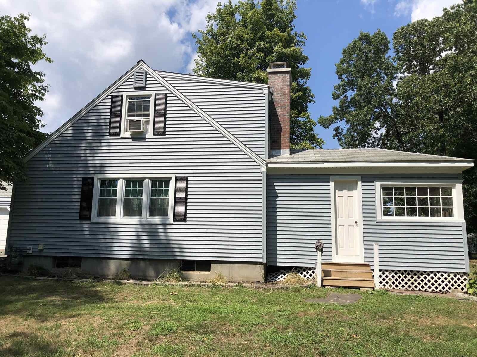 A two-story grey, vinyl-sided house with a white door, chimney, and large windows set against a blue sky with green trees.