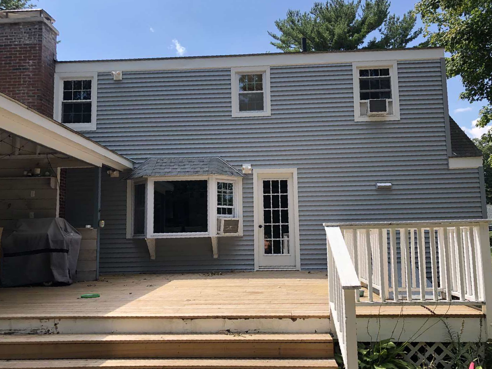 A two-story blue house with light-colored siding, a wooden deck, a bay window, and a white back door under a sunny sky.