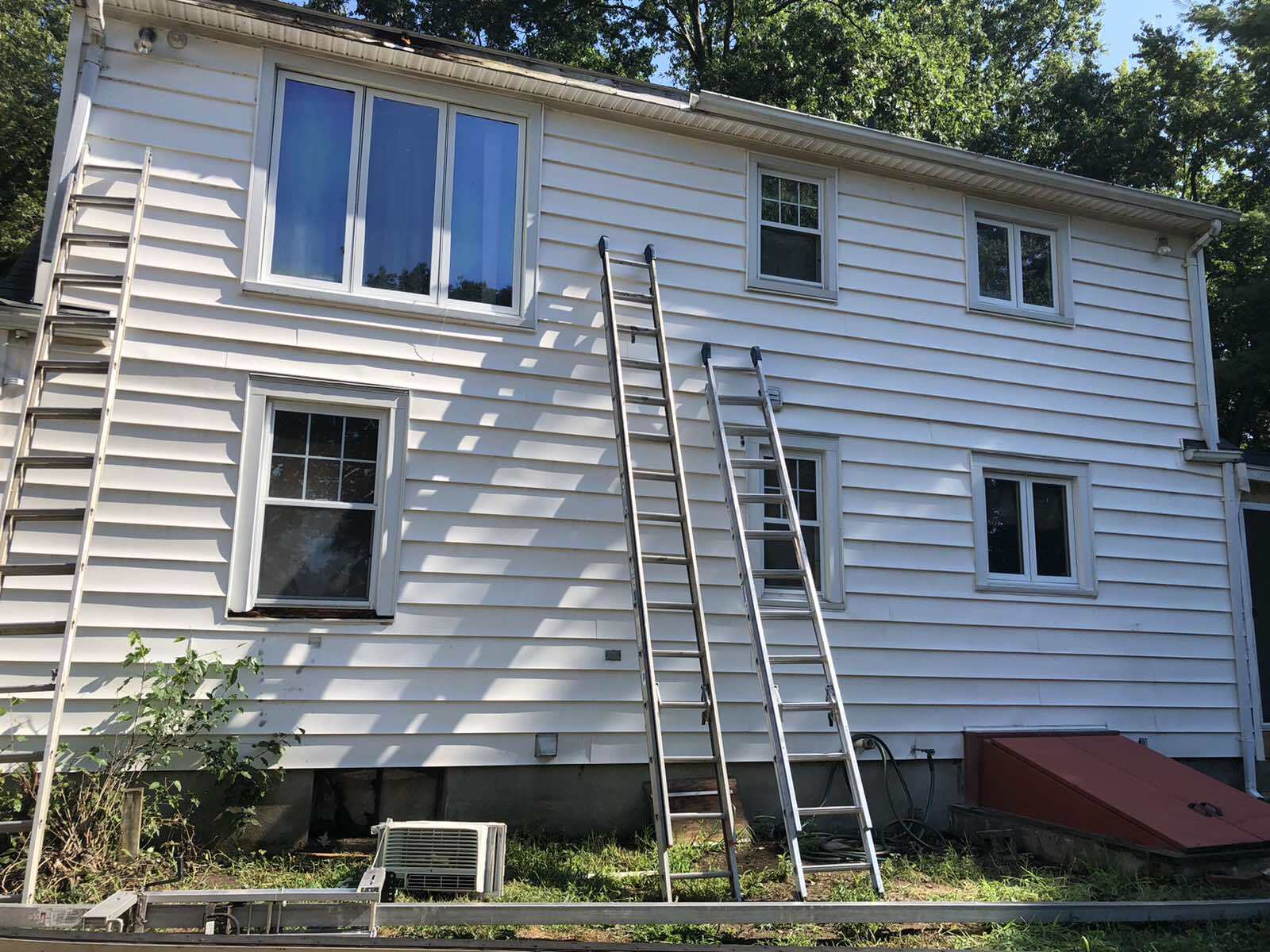 A two-story white house with siding, three windows, and two extension ladders leaning against the exterior wall.