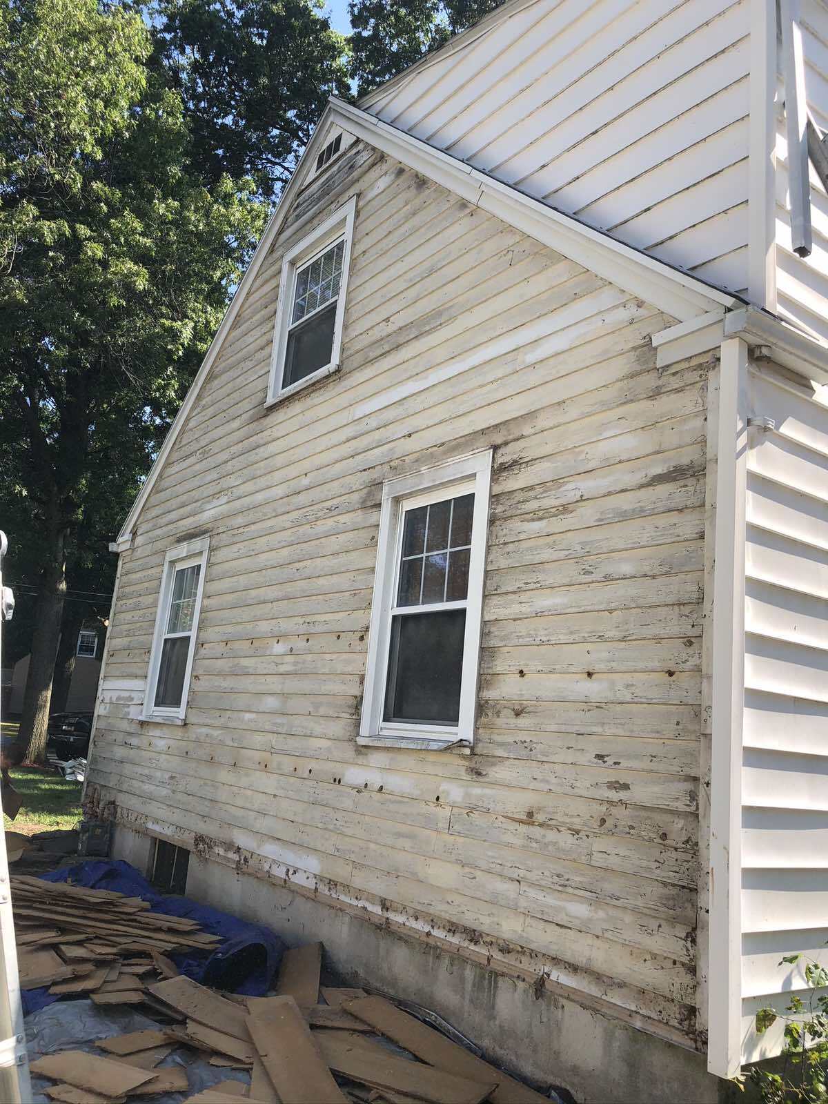 The exterior of a house with light-colored siding removed, exposing the wall sheathing, with debris on the ground.