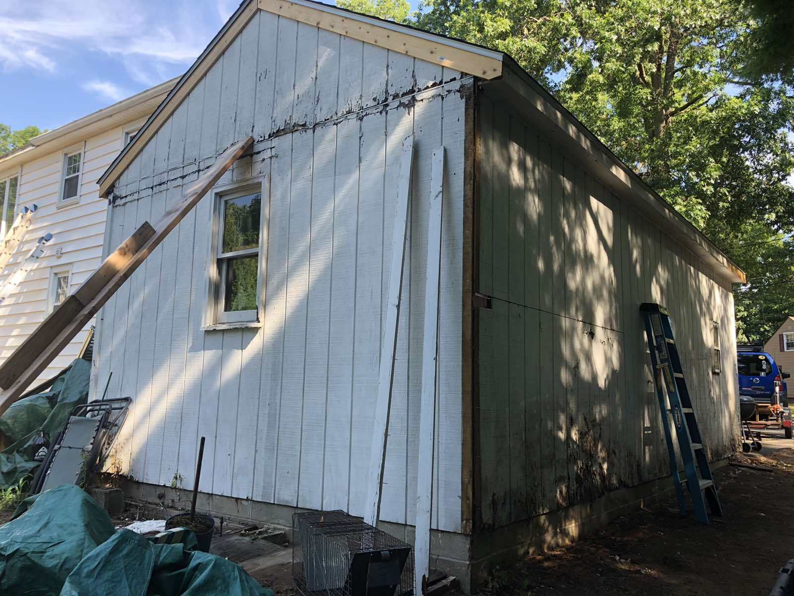 A side view of a white, vertical-sided shed under construction, showing a window, an attached ladder, and exposed wood.