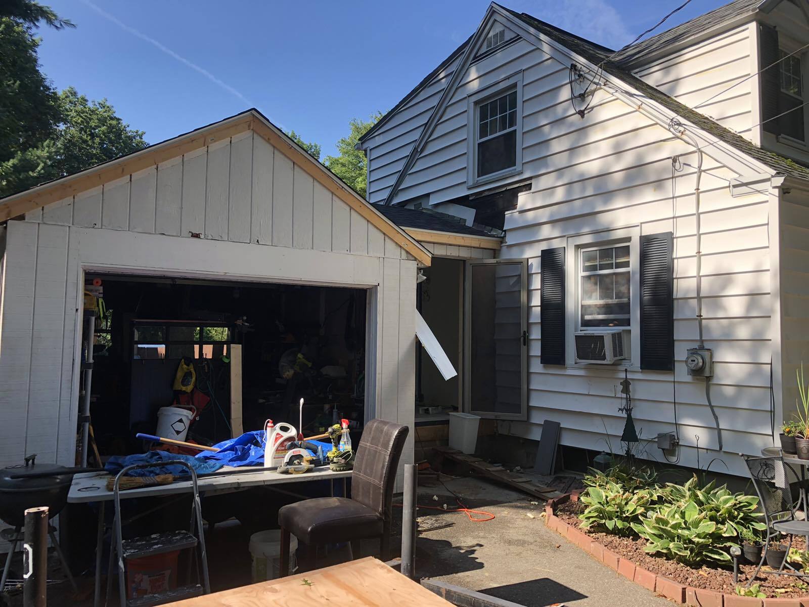 A house and a detached garage with an open door, facing a concrete patio with a table, chairs, and plants.