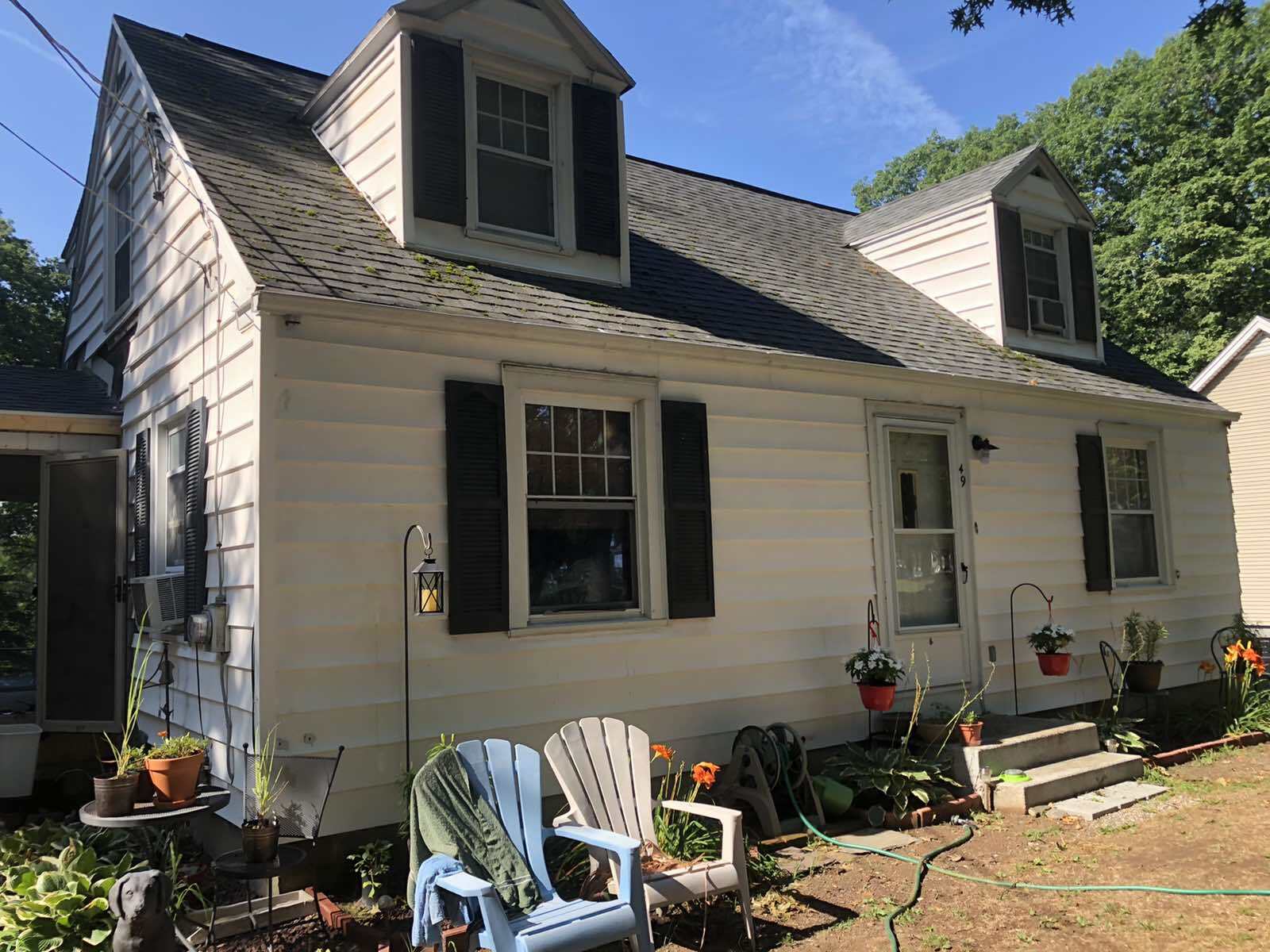 A white, one-and-a-half-story house with two dormers, black shutters, and blue and white chairs on the lawn.