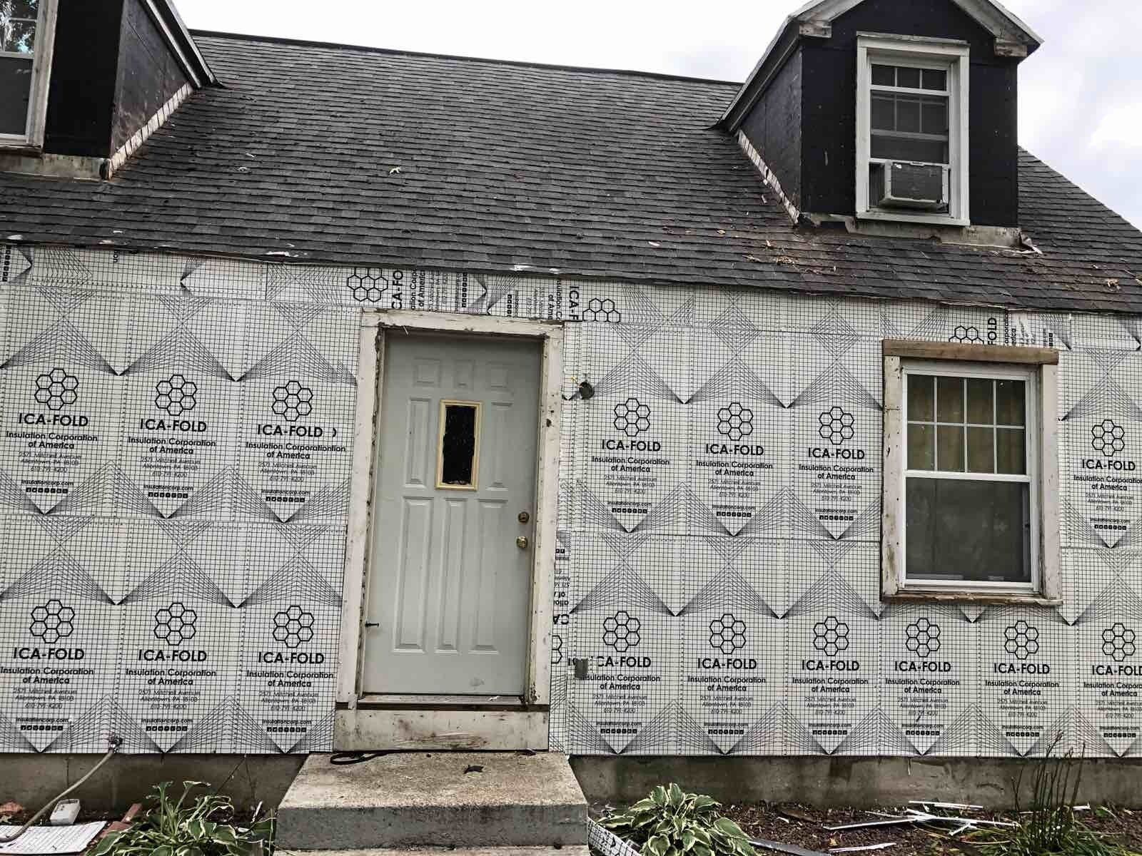 A light-colored house with unfinished insulation siding, a centered door, a window on the right, and two roof dormers.