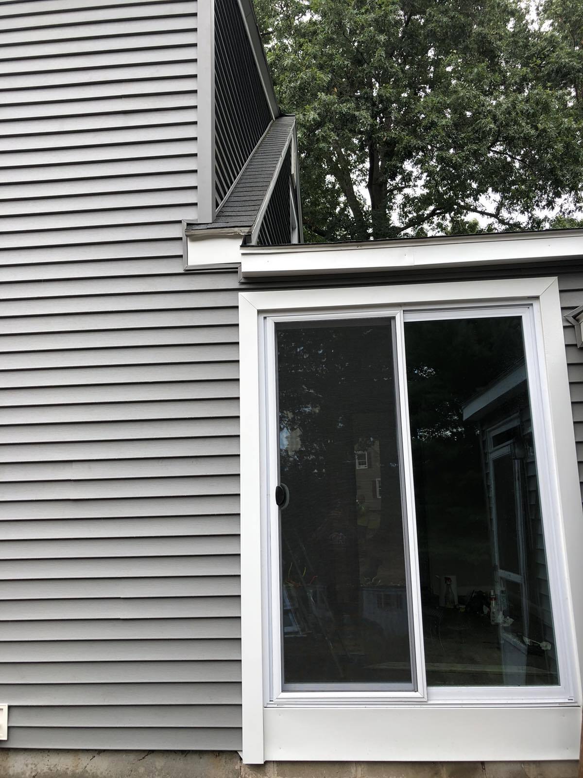 A white sliding glass door set into the gray vinyl siding of a house, beneath a roof corner and near mature trees.