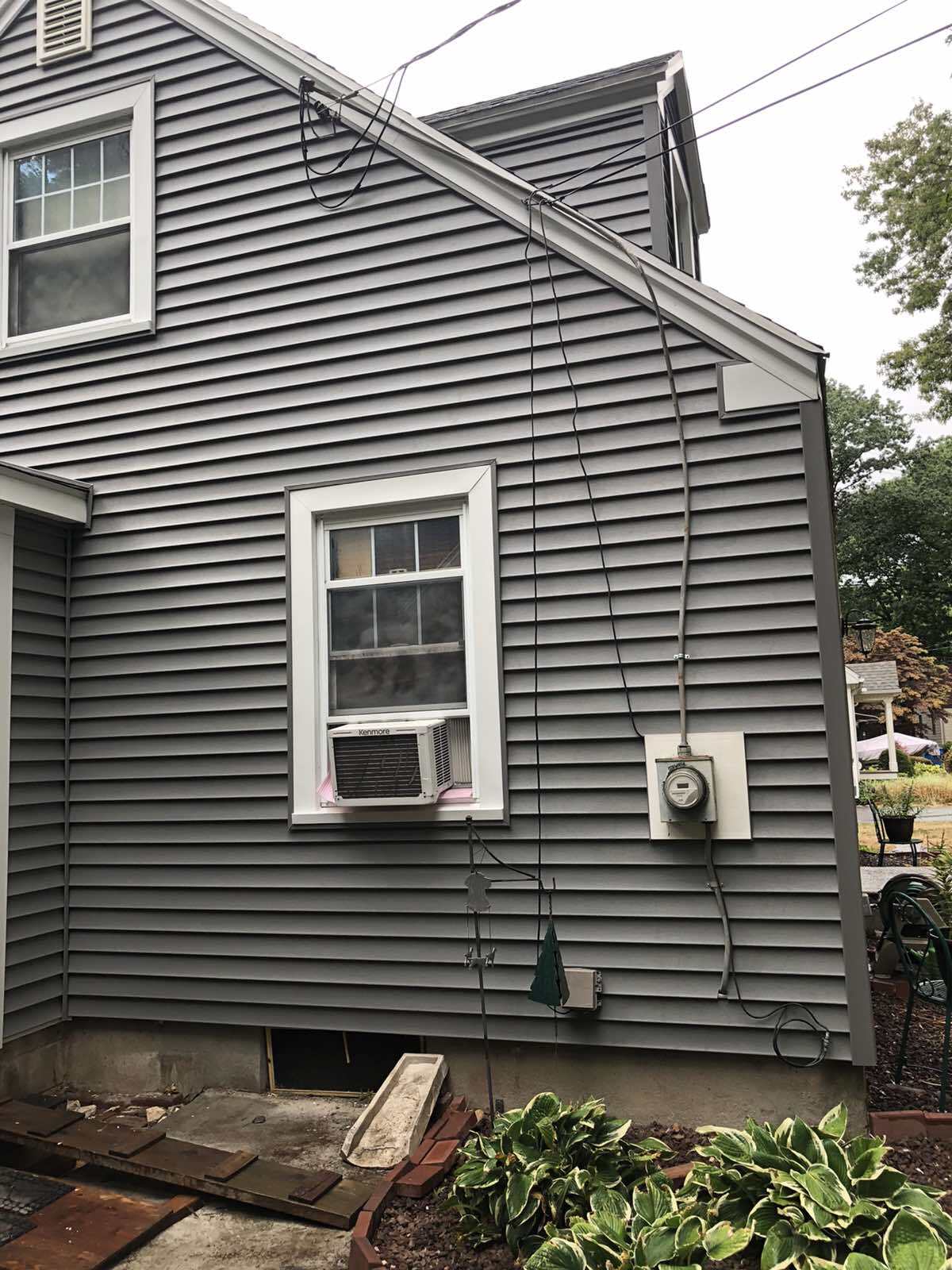 A side view of a gray-sided house exterior featuring a window-mounted air conditioner and an electrical meter on the wall.