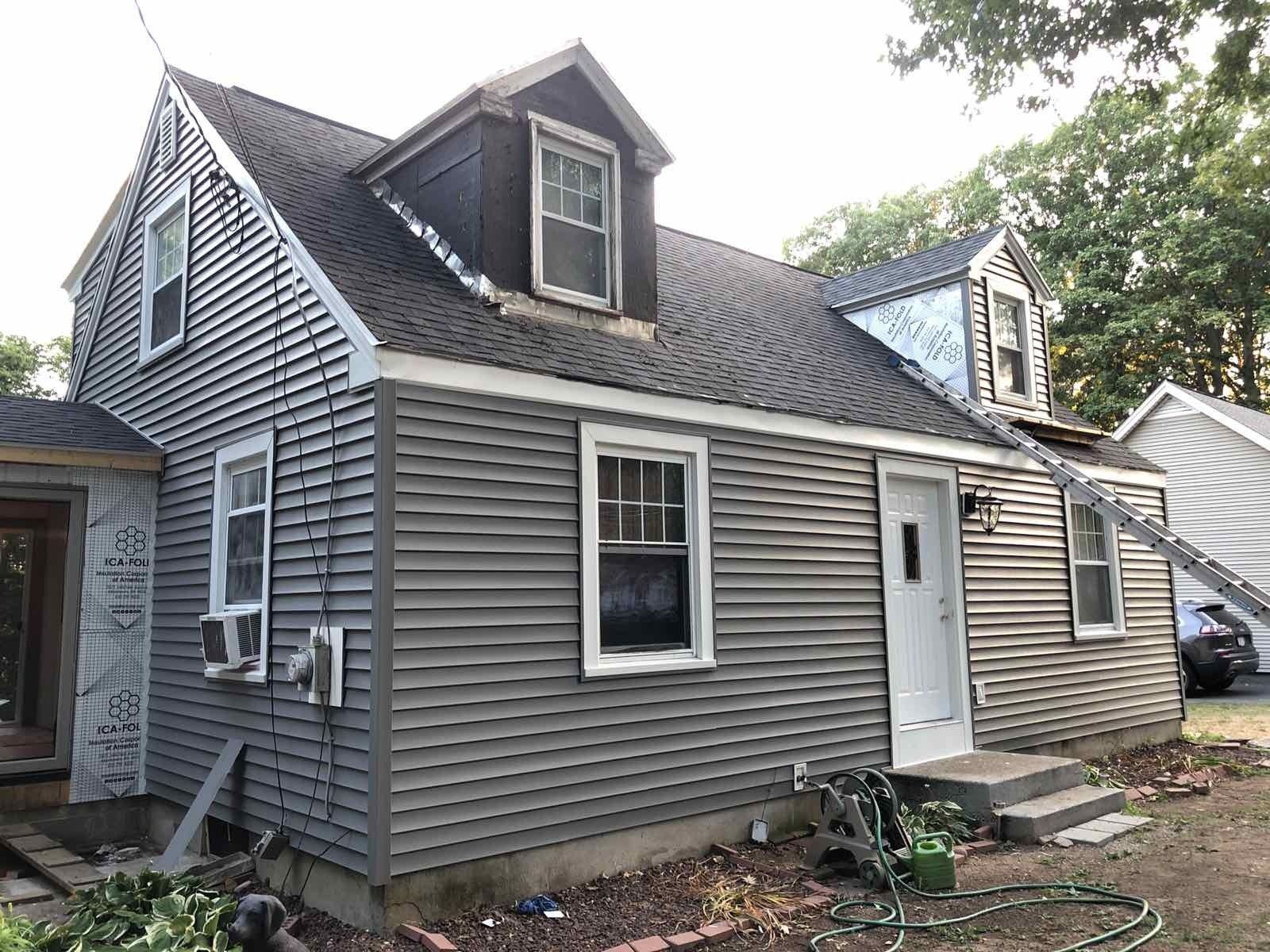 A grey two-story house with two dormer windows and ongoing exterior renovations, seen from an angled outdoor perspective.