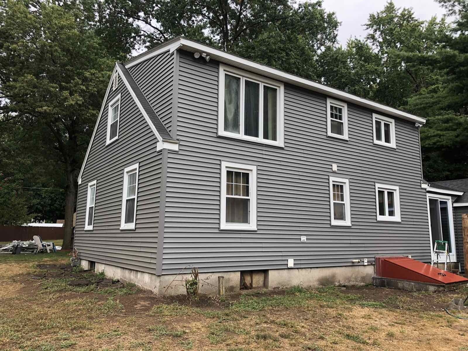 A two-story house with grey vinyl siding, white window trim, a concrete foundation, and a small red bulkhead door.