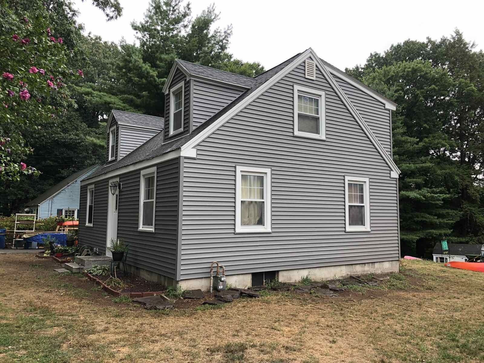 A gray-sided, two-story house with dormer windows, white trim, and a yard with trees in the background.