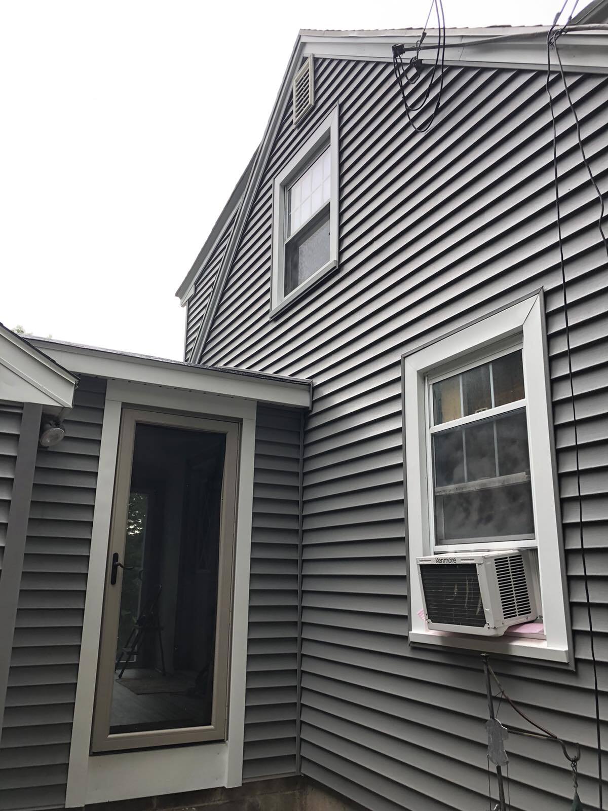 A grey, two-story house exterior with a door and a window containing a white air conditioning unit.