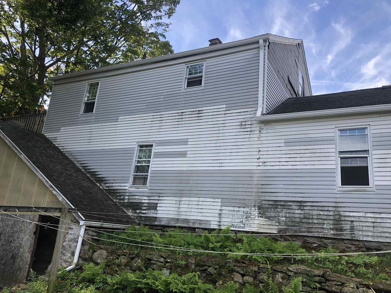 A two-story house with gray and white siding needing repair, situated above a stone wall and surrounded by greenery.
