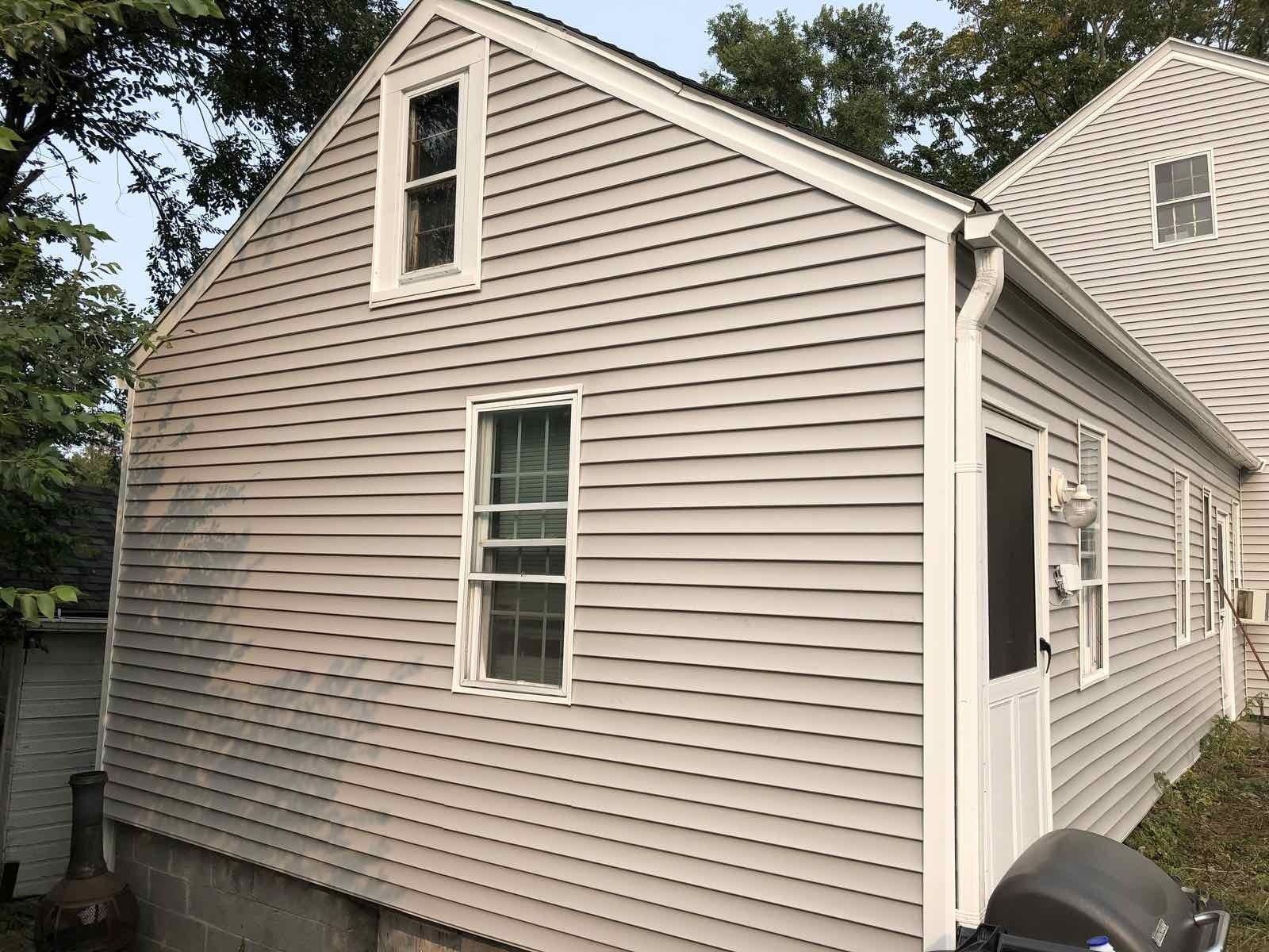 The side and back of a light-gray house with vinyl siding, two windows, and a white-framed screen door.