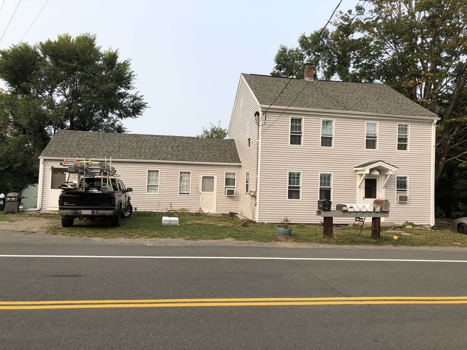 A two-story white house with an attached single-story section and a pickup truck parked in front, beside a two-lane road.
