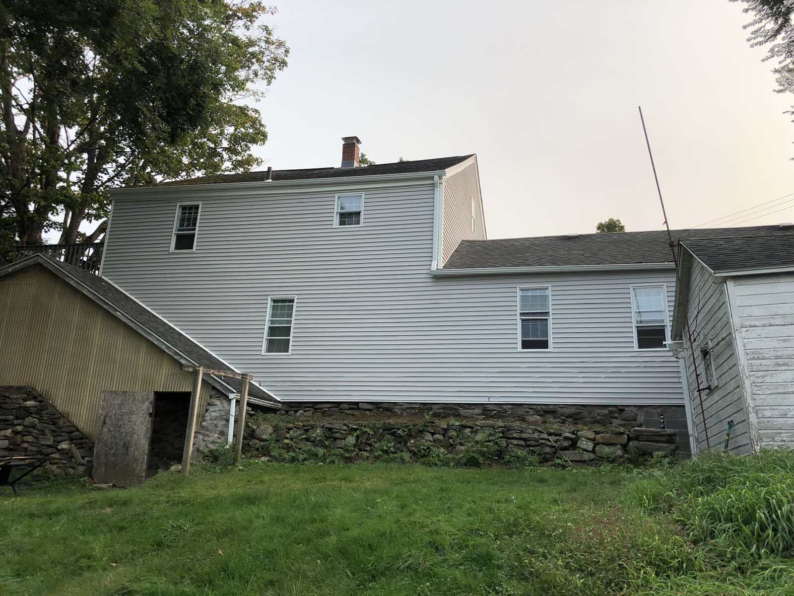 A two-story house with light-colored horizontal siding, a stone foundation, and a small wooden shed extension.