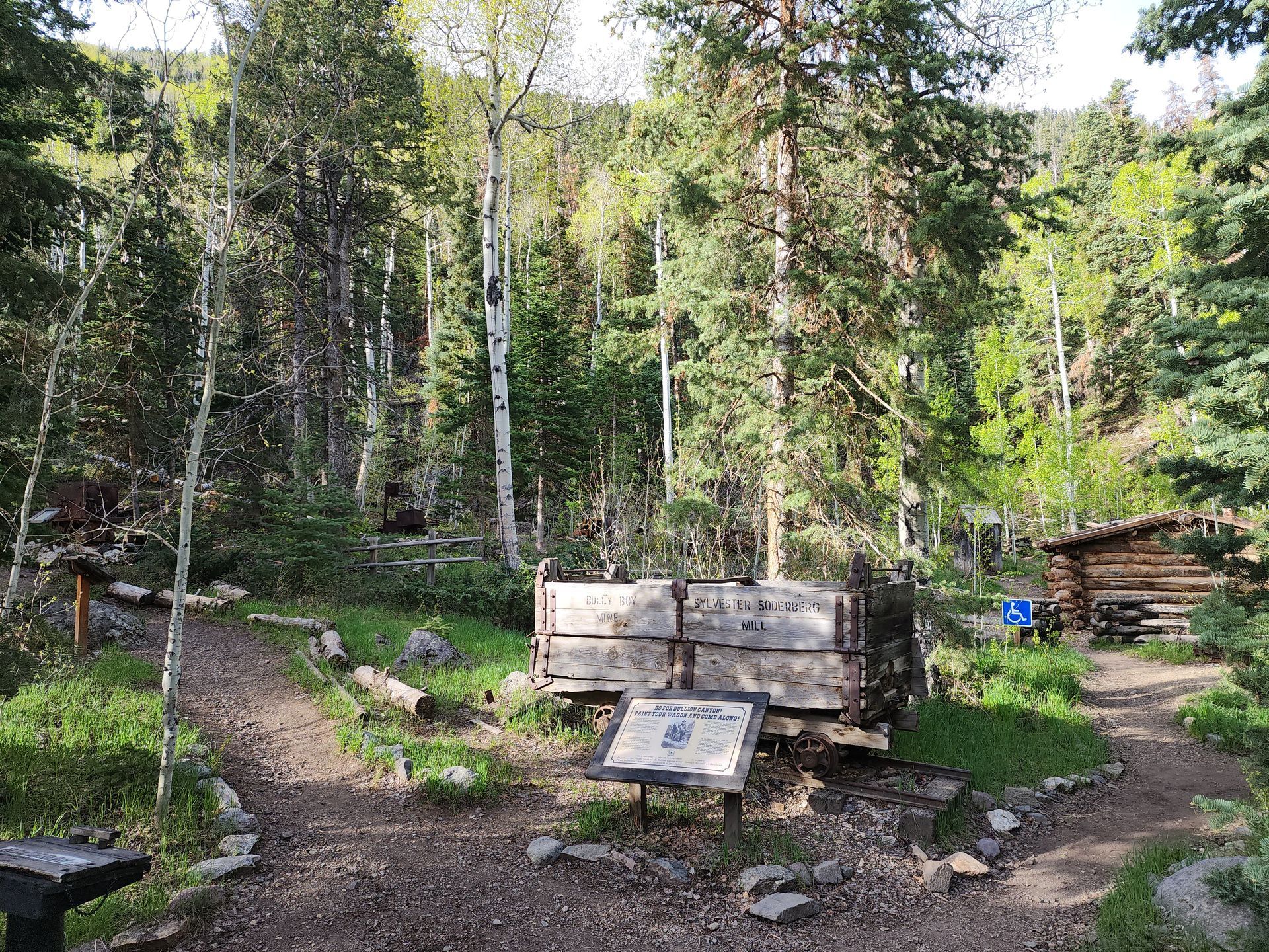 A weathered mining cart sits on a dirt path in a wooded area near a small log cabin, with a sign in front.
