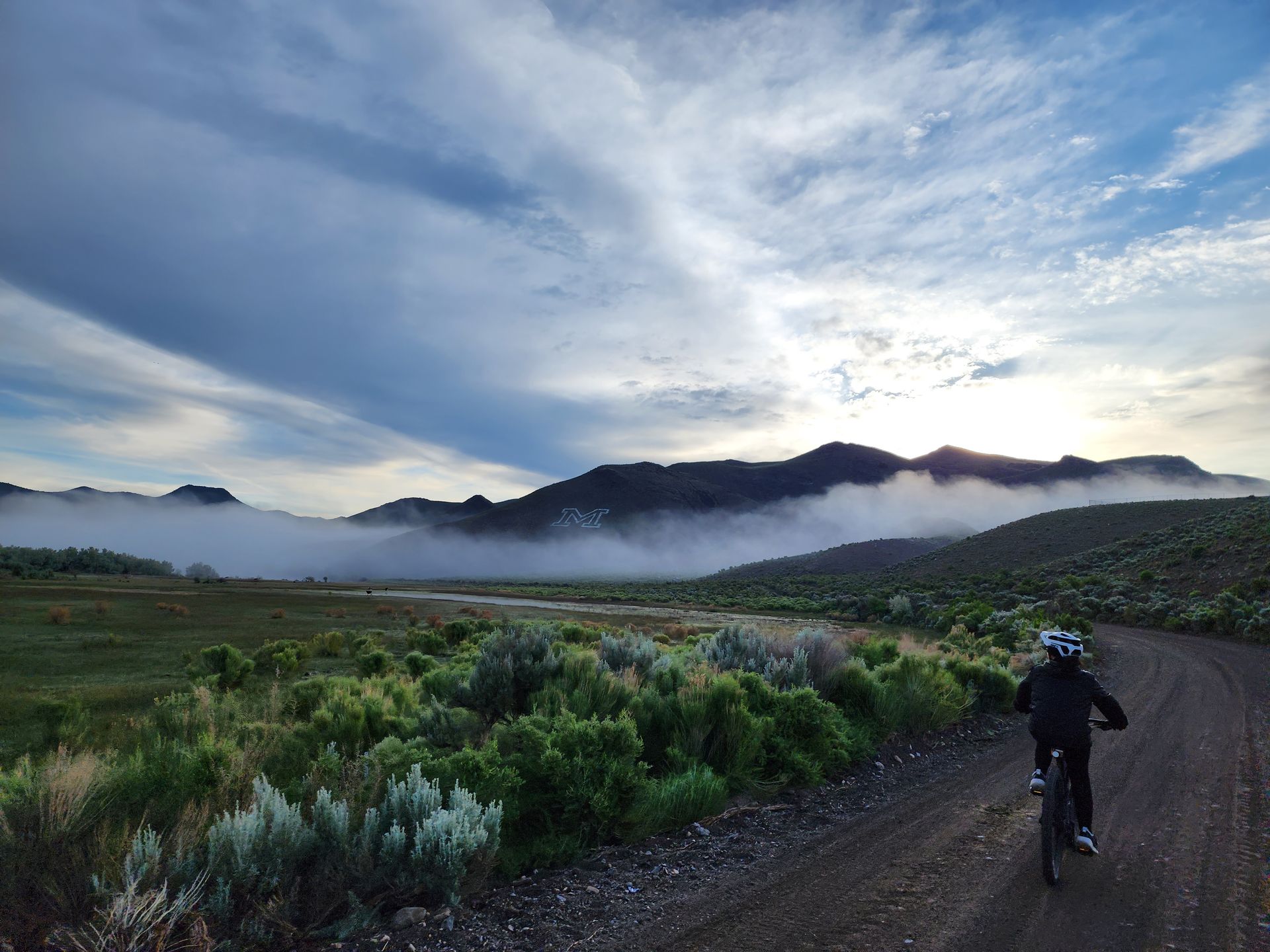 A cyclist rides a dirt path toward mountains partially obscured by low-hanging clouds under a partly cloudy sky.
