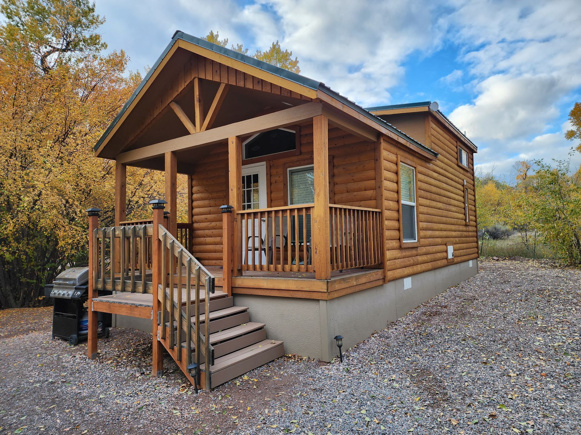 A small, brown log cabin with a covered front porch, wooden stairs, and a grey foundation set on a gravel lot.