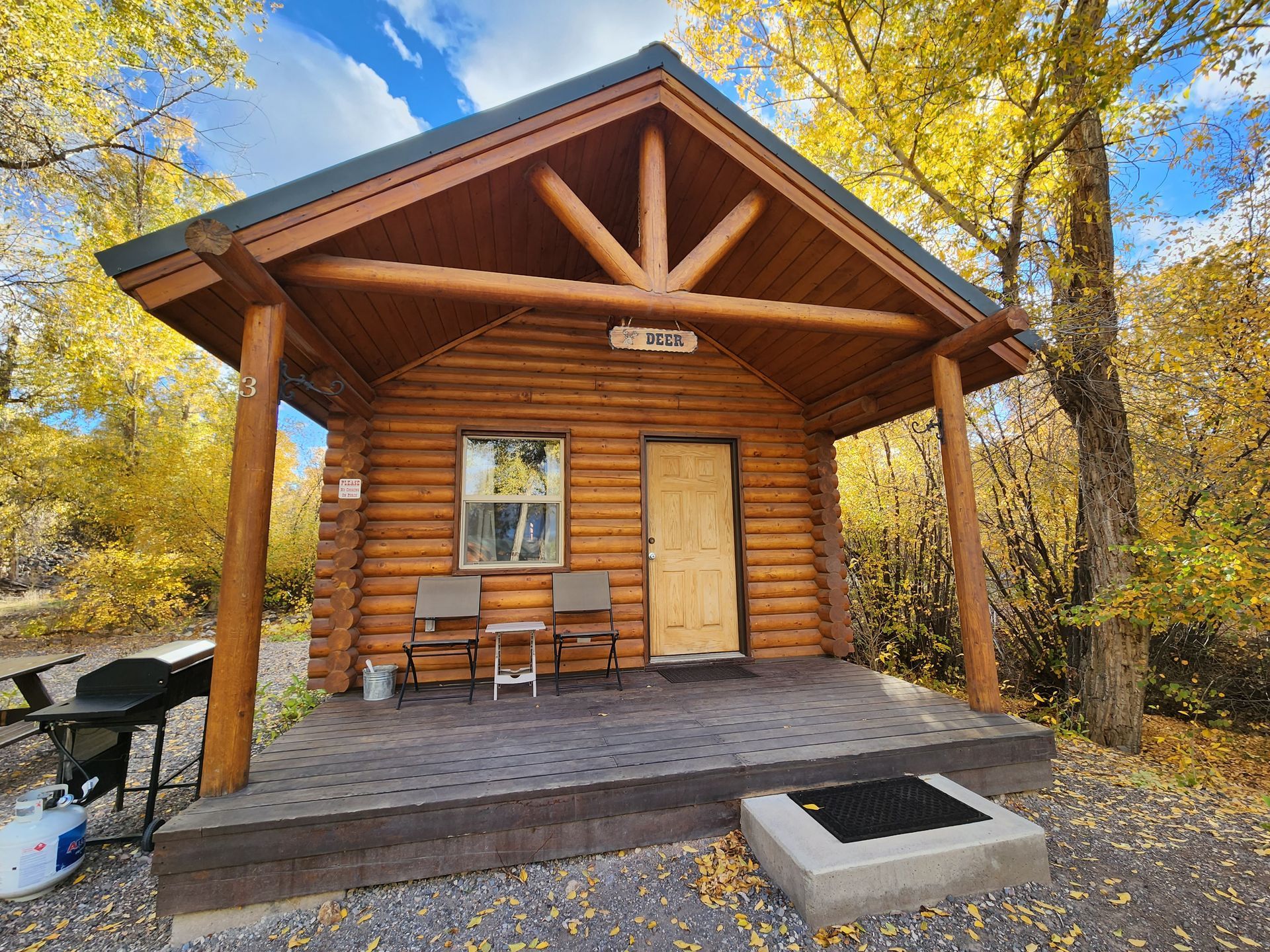 A small log cabin with a front porch, two chairs, and a grill, surrounded by trees with yellow autumn leaves.