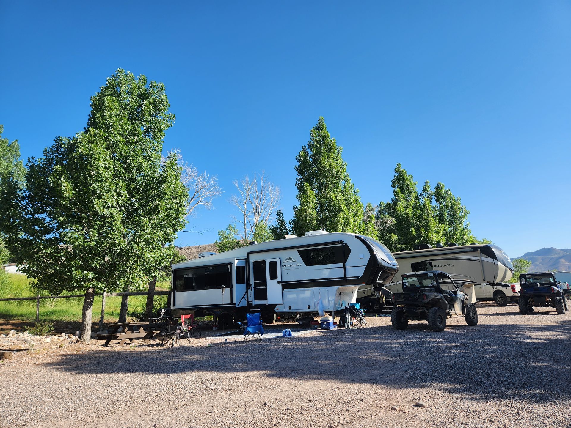 Two RVs and two off-road vehicles parked on a gravel lot under large trees on a sunny day with a clear blue sky.