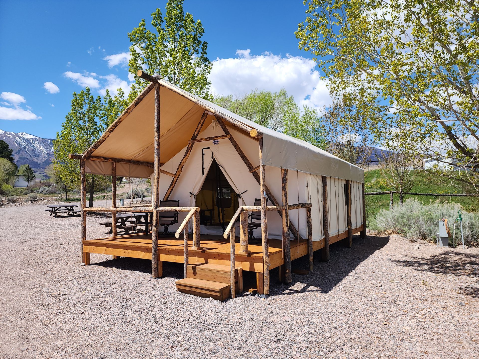 A canvas glamping tent on a wooden deck with a covered porch, set on a gravel lot near trees and snowy mountains.