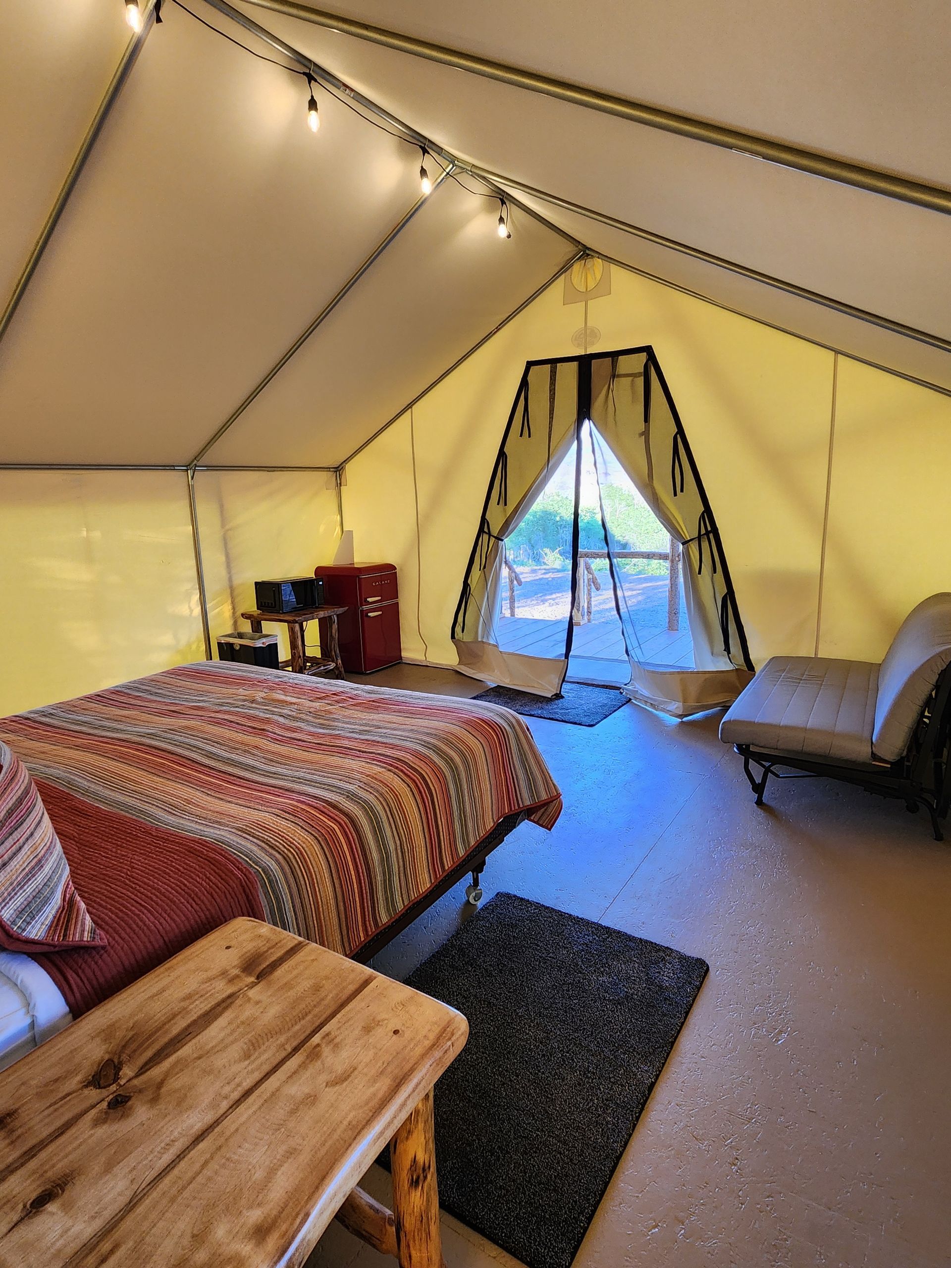 Inside a tent with a bed, small wooden table, rug, and folding chair, looking toward an open entrance.