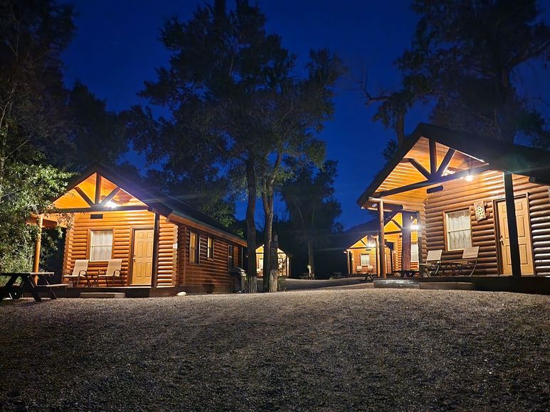 A rustic log cabin with a wooden porch, two chairs, and a metal roof, set in a wooded outdoor area.