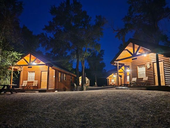 Autumnal trees with golden leaves surround wooden cabins and parked vehicles under a clear blue sky.