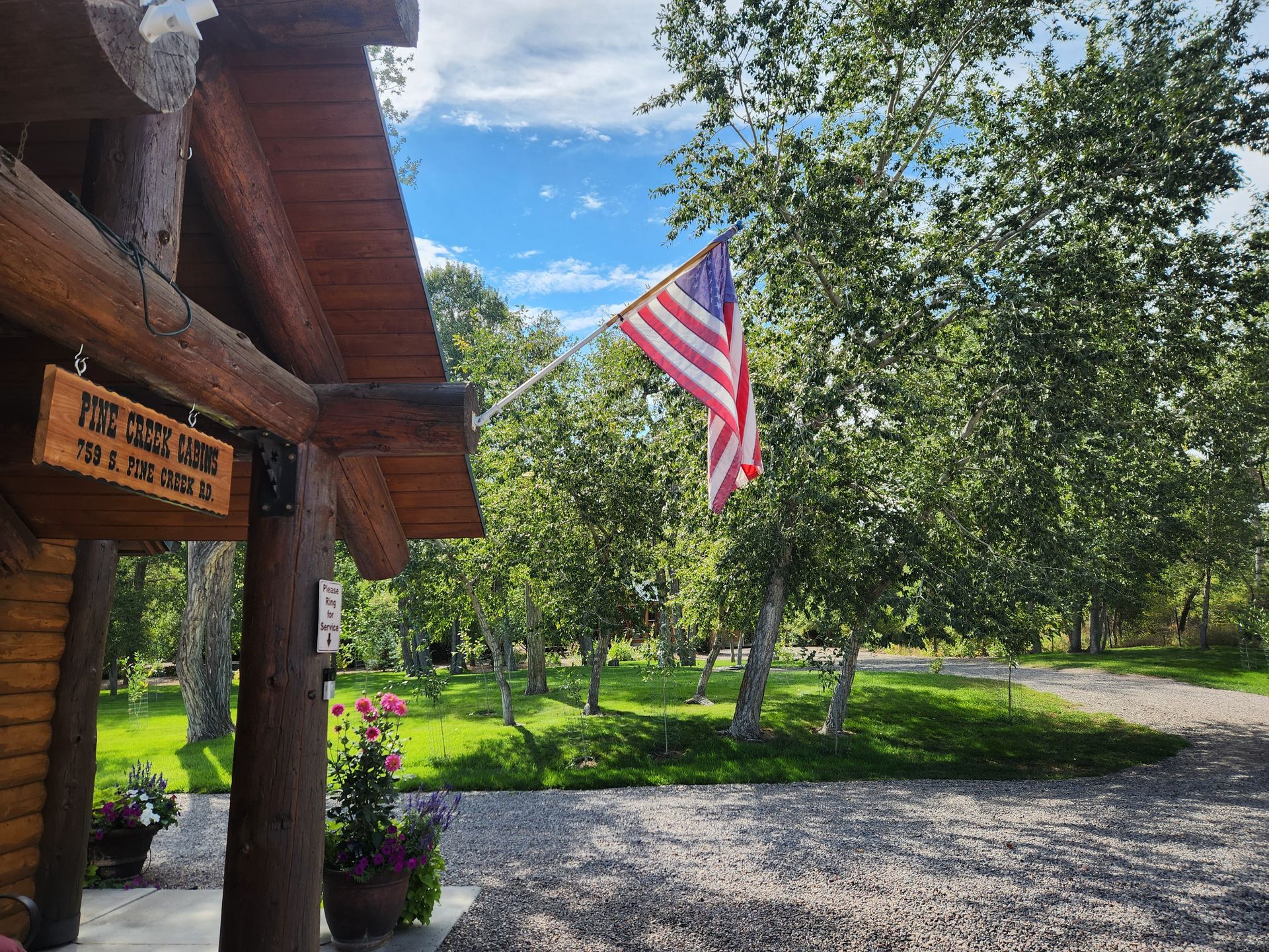 A wooden building with an American flag flying, overlooking a gravel path, green trees, and a grassy area.