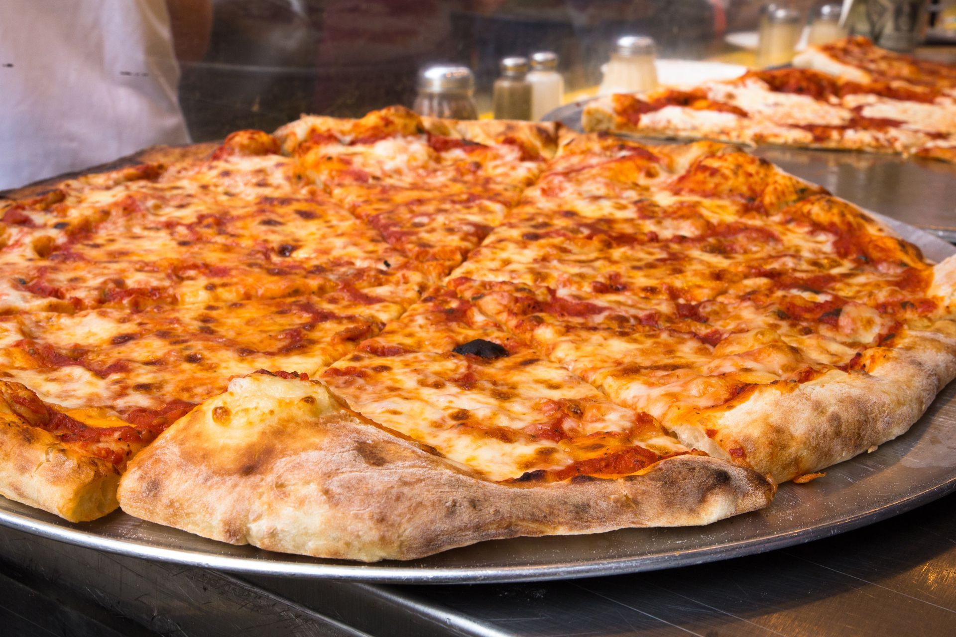 A cheese pizza on a metal tray sits on a counter, with another pizza visible in the background.