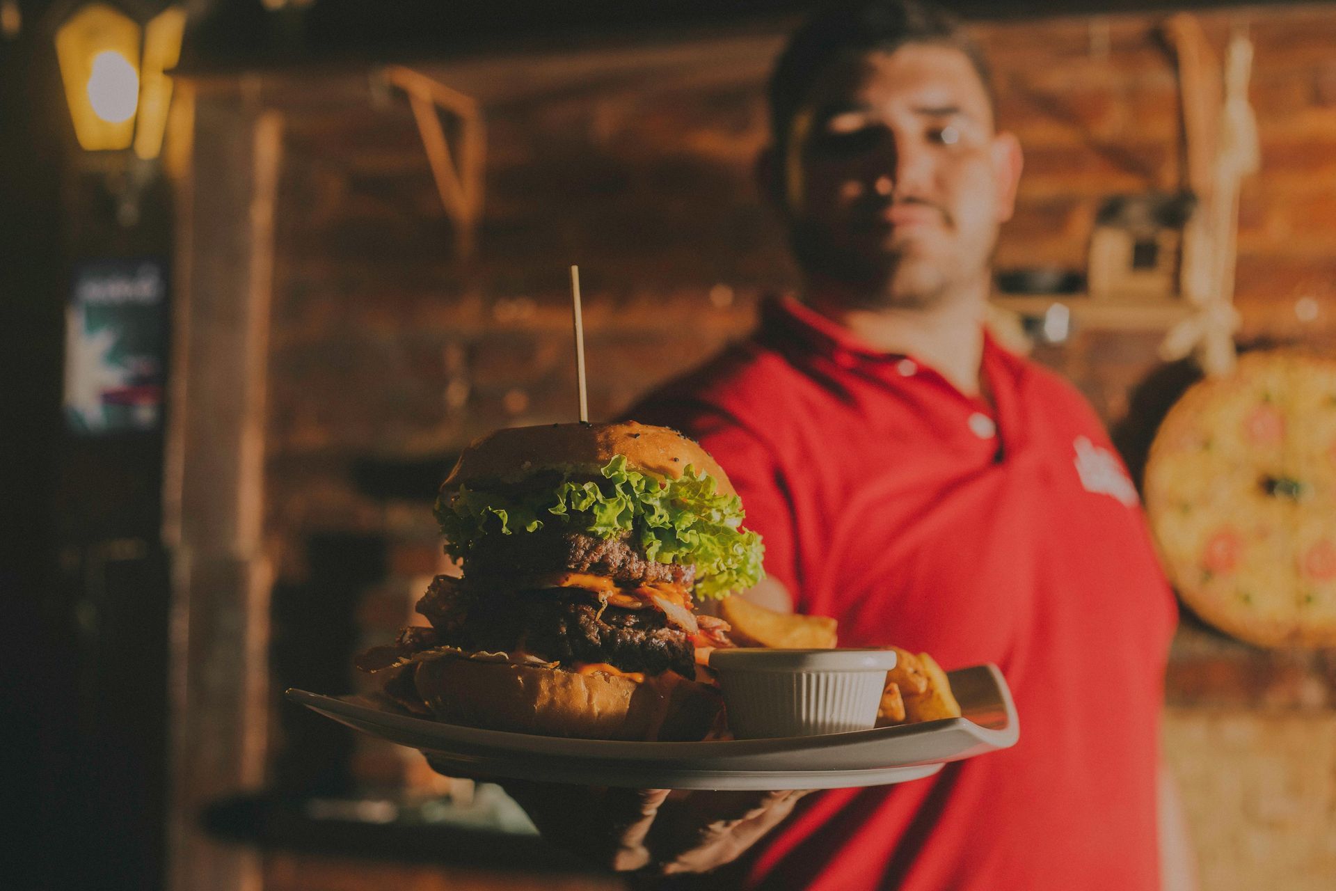 A person in a red polo shirt holding a large burger and a small dish of sauce on a plate in a dimly lit, rustic space.
