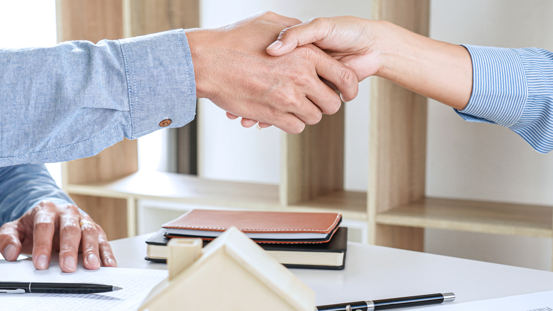 Two people shaking hands at a desk. A small house model and documents are visible.