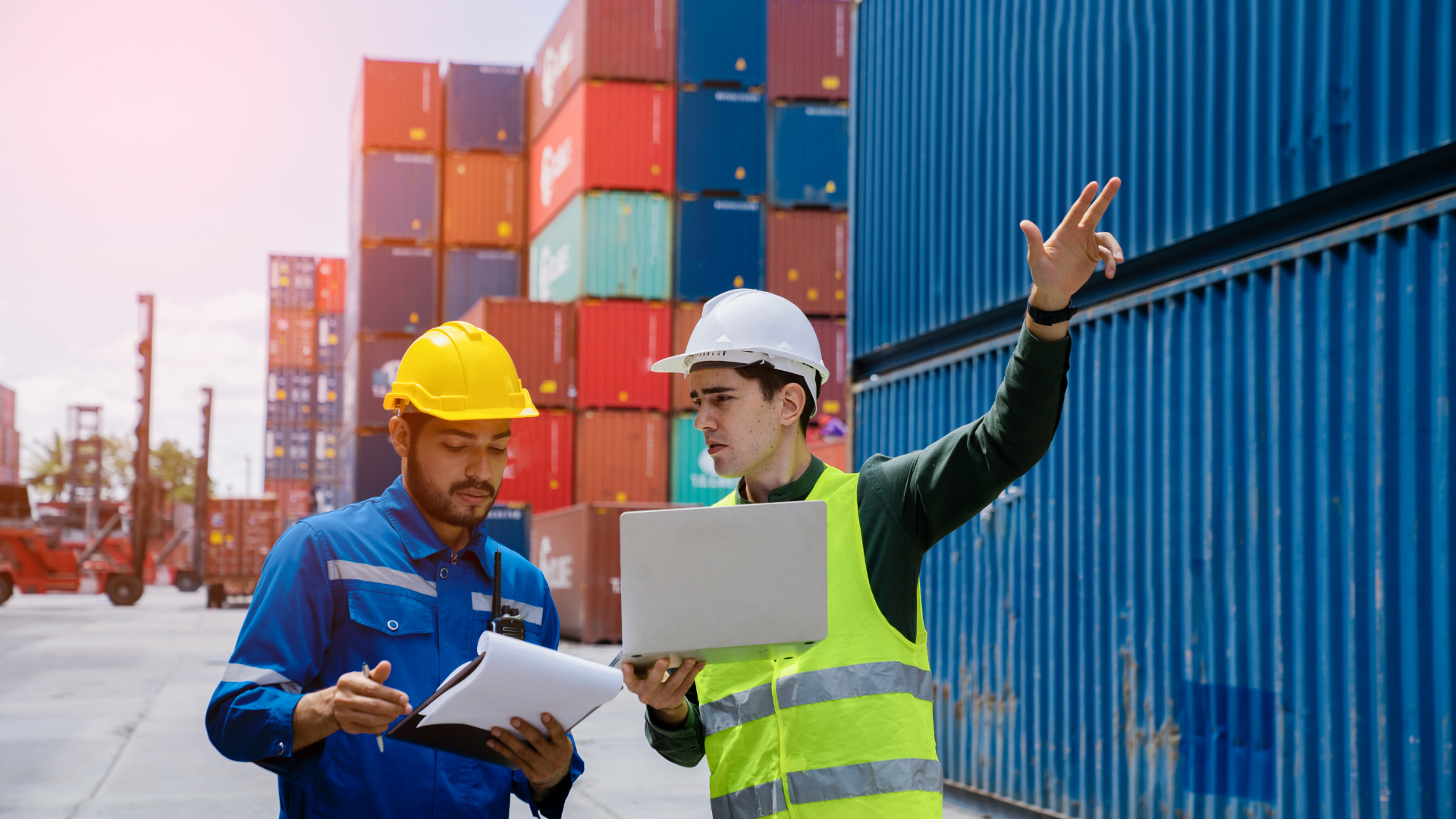Two men in safety vests and hard hats inspect cargo containers at a port.
