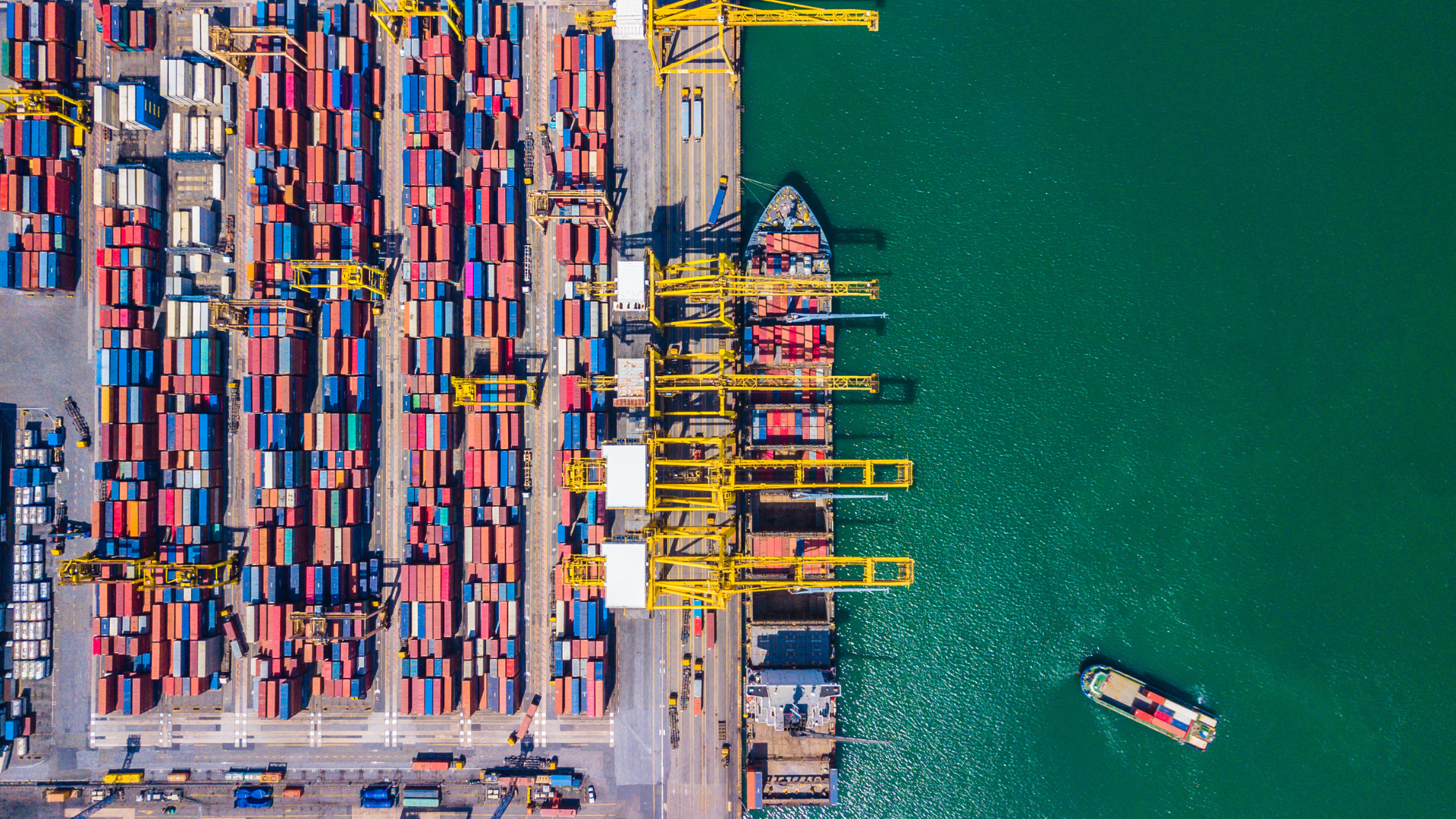 Aerial view of a busy shipping port; cargo containers, ships, cranes, and small boat on turquoise water.
