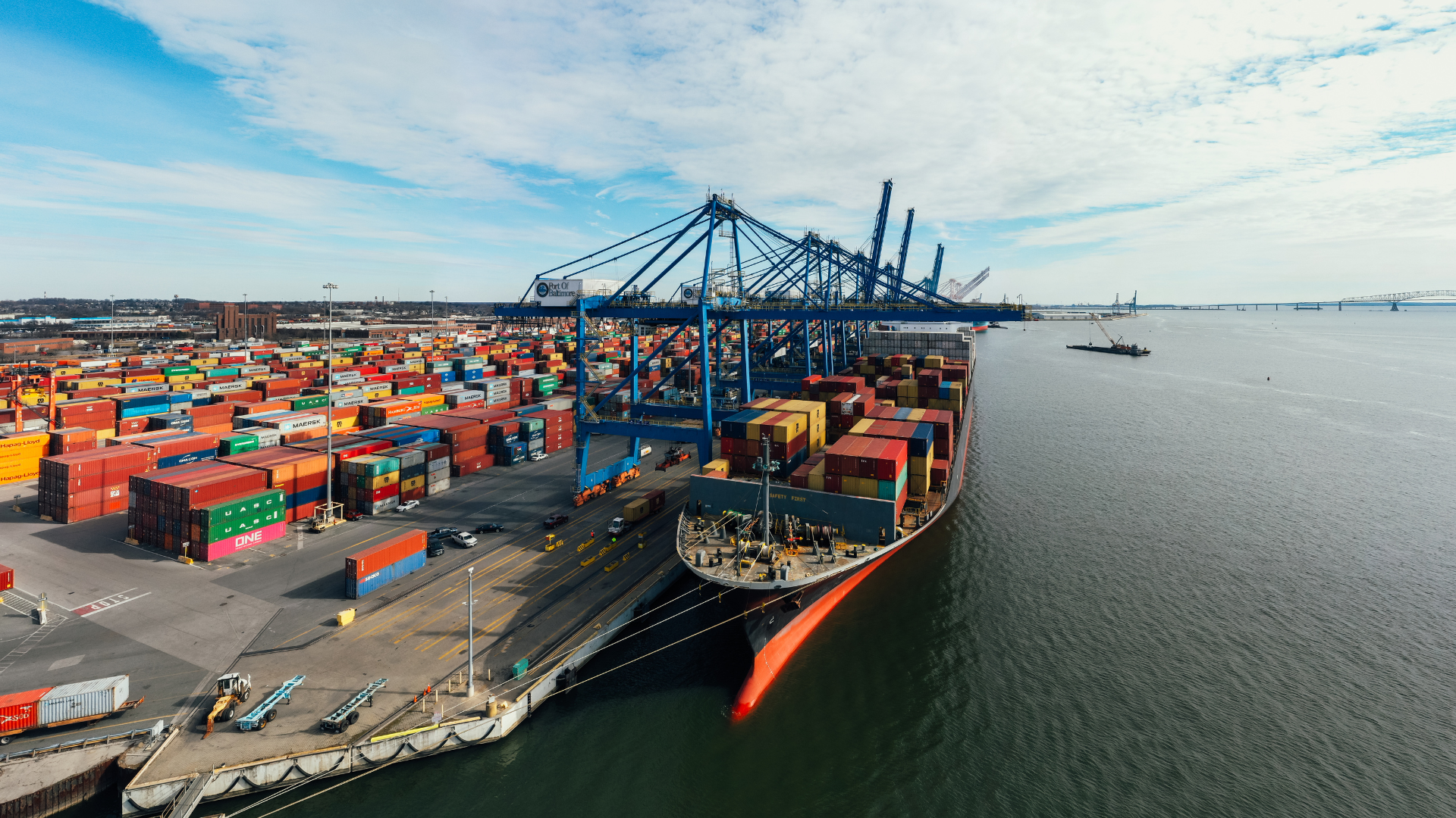 Cargo ship docked at a port, being unloaded by large cranes. Containers are stacked on the dock.