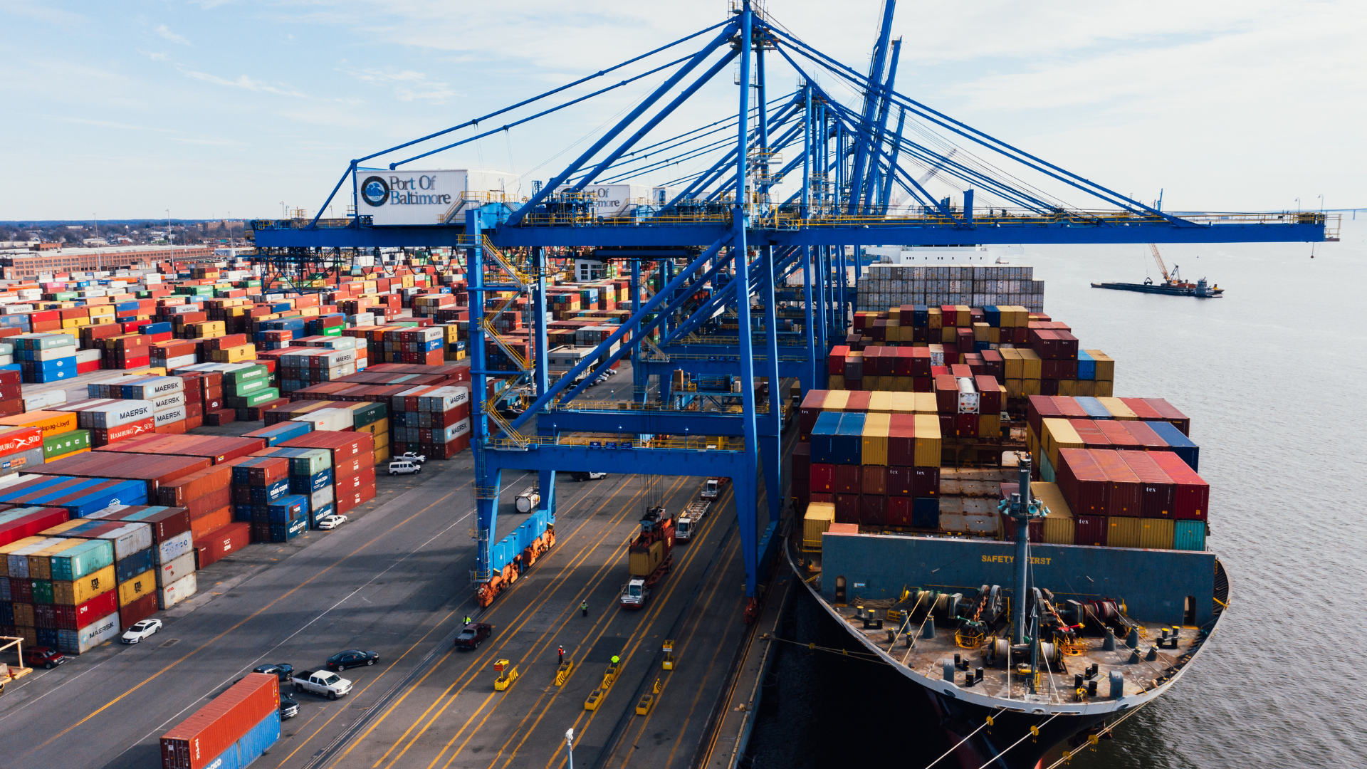 A cargo ship docked at a port, being unloaded by blue cranes, surrounded by containers.