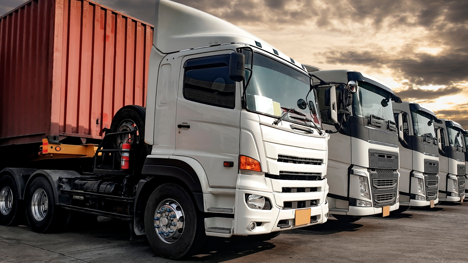 Semi-trucks lined up, white truck in focus, orange container on the left, parked outdoors against a cloudy sky.