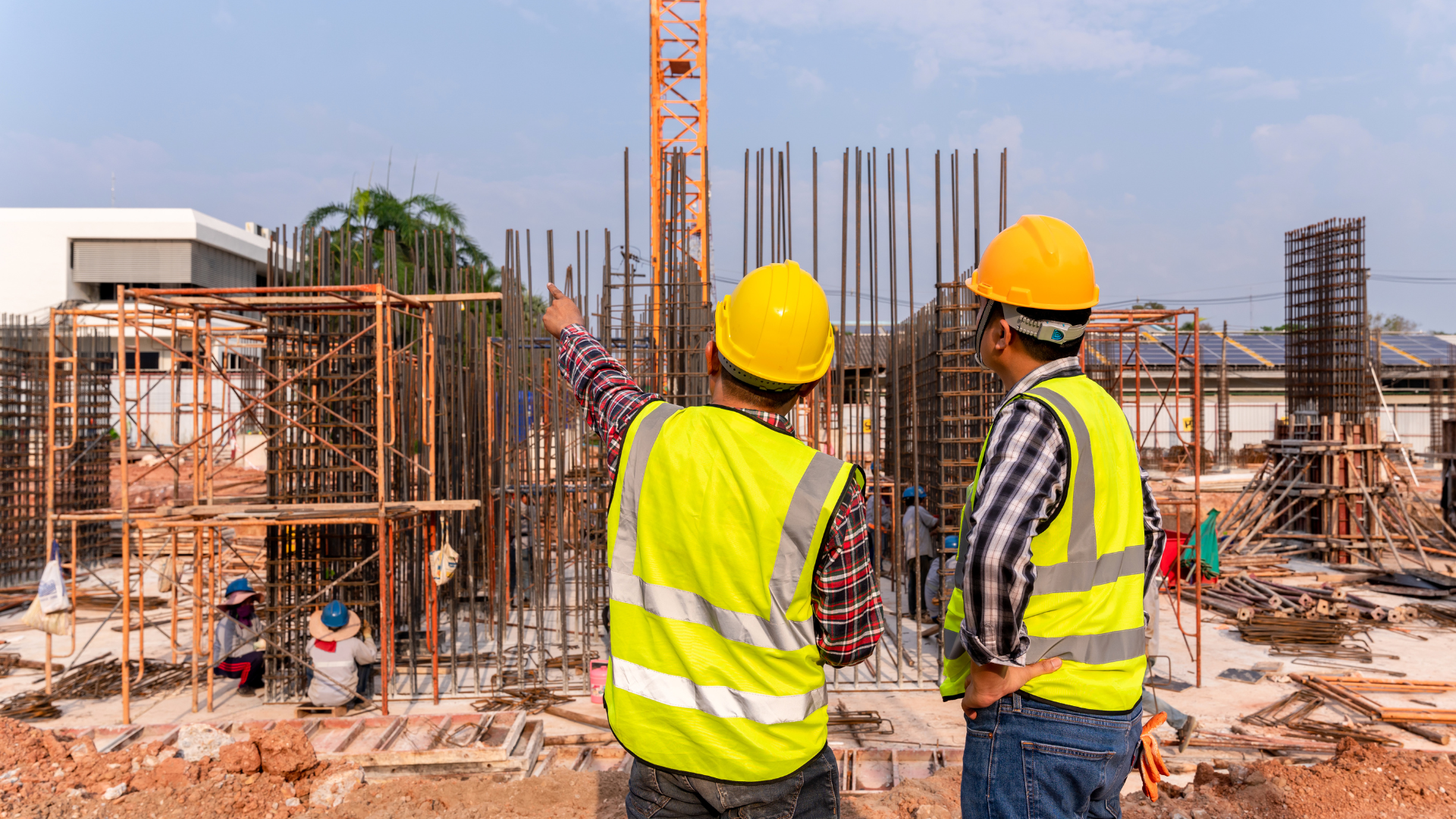 Two construction workers in yellow vests and hard hats at a building site, one pointing upwards.