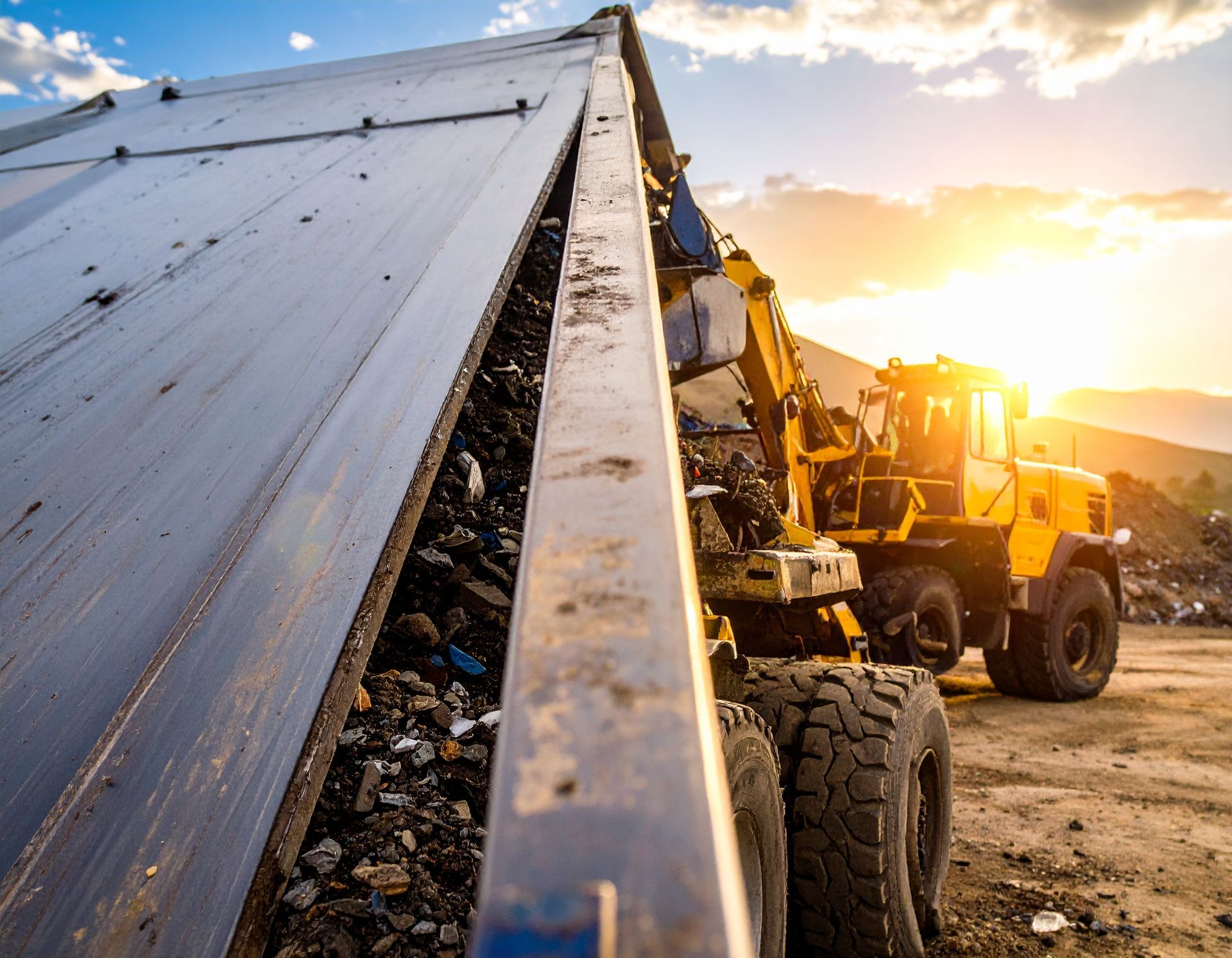 A yellow dump truck unloading trash with a setting sun in the background.