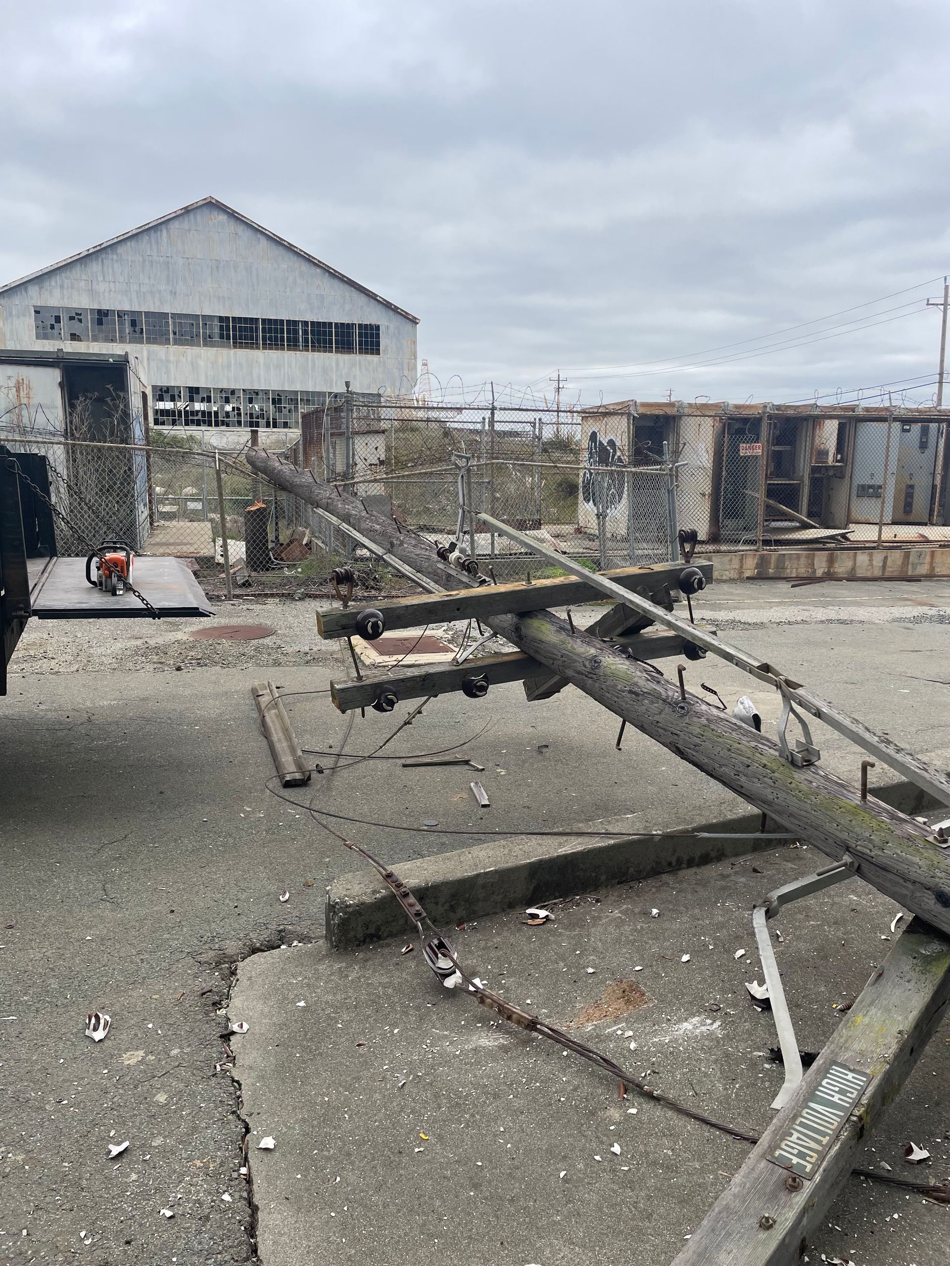 A downed power pole on a cracked concrete surface; industrial buildings in the background under a cloudy sky.